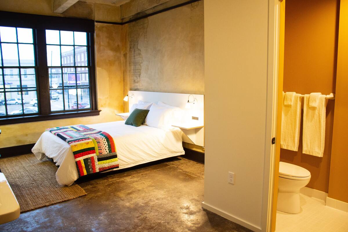 A bedroom with a large window, a bed with a colorful quilt, and a view of the street outside.