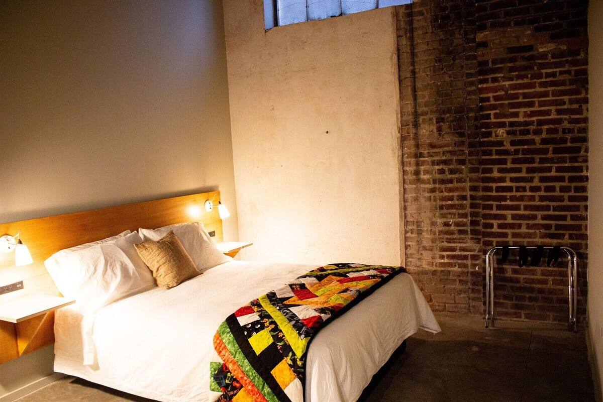 A hotel room with a bed, a nightstand, a wall-mounted lamp, a brick wall, and a metal radiator.