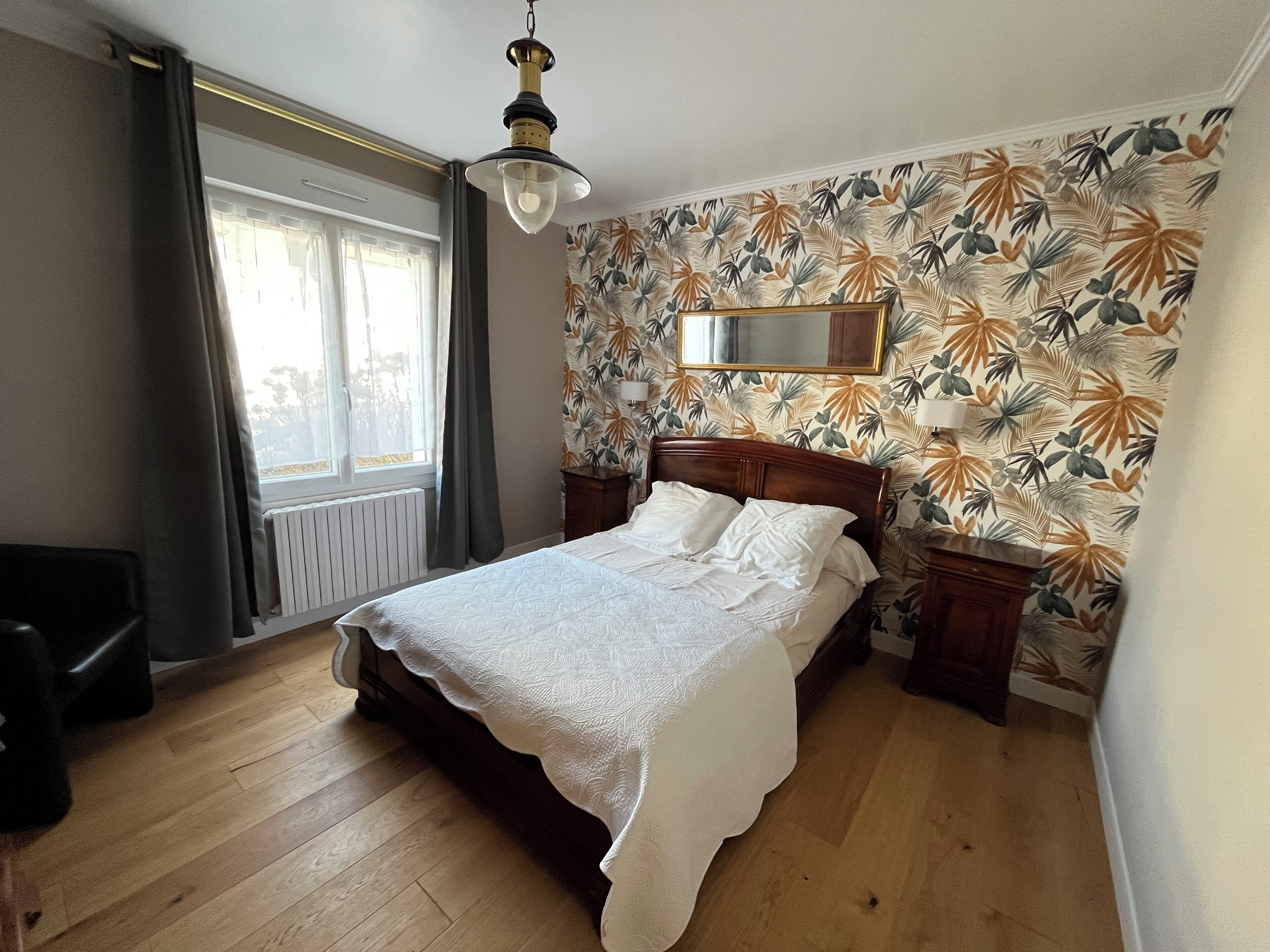 A bedroom with a wooden bed, a window with curtains, a radiator, and a wall with a tropical leaf pattern.