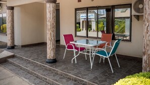 Un espacio al aire libre techado con mesa y sillas, una puerta de vidrio que da a un estacionamiento y un ventanal con vista al área de estacionamiento.