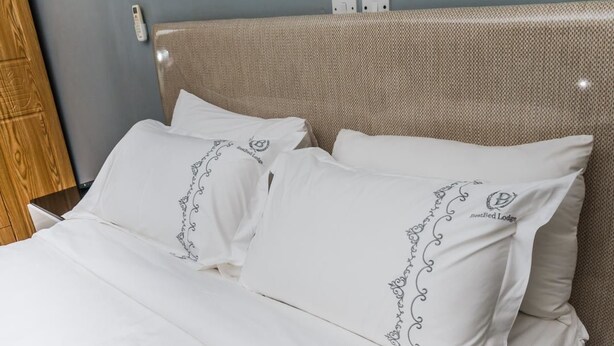 Una cama con ropa de cama blanca y almohadas decorativas, una mesita de noche de madera y un interruptor de luz en la pared.