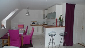 Cocina compacta con gabinetes blancos, microondas y cafetera. Hay un comedor con sillas rosadas y una mesa.