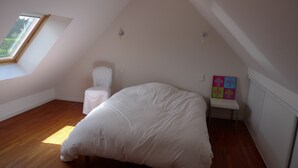 Un dormitorio con techo inclinado, una cama individual, una silla, una mesita con un cuadro colorido y una ventana con vistas a zonas verdes.