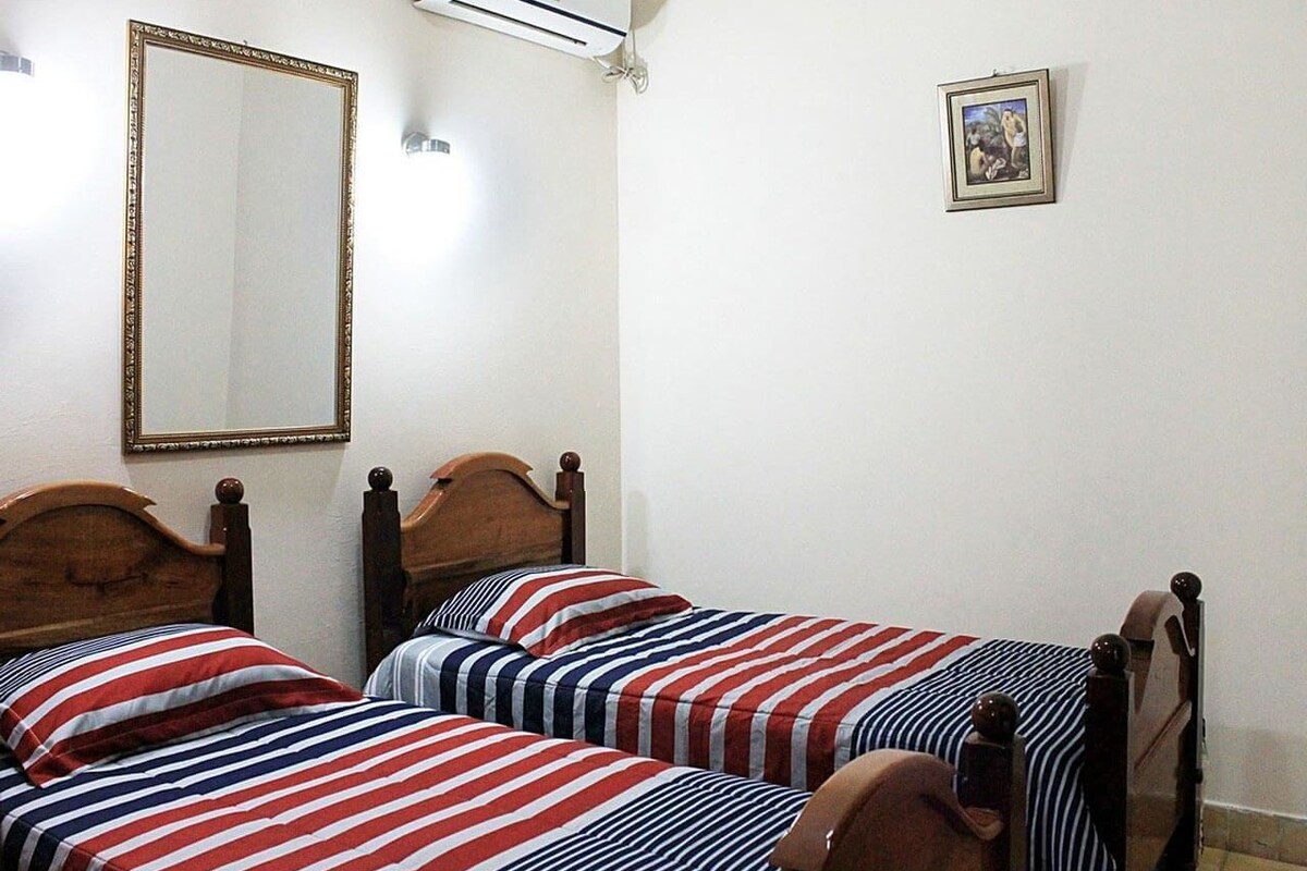 A room with two wooden beds with striped bedding, a wall-mounted air conditioner, and a framed picture on the wall.