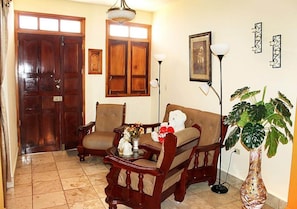 A living room with a wooden sofa, armchairs, a coffee table with decorative items, a potted plant, and a large wooden door. - Hostal Merlyn TRINIDAD (Trinidad)