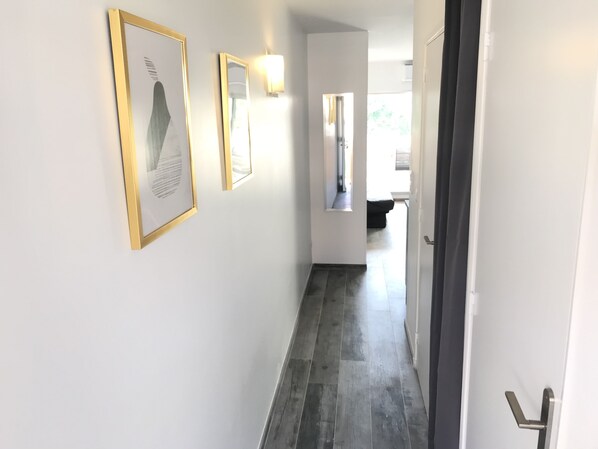 A hallway with a dark wooden floor, white walls, and framed artwork. There is a door on the right and a window at the end of the hallway. - Apartment renovated, air conditioning, sea view, swimming pools, tennis (Antibes)
