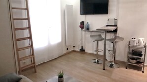 Moderni, pelkistetty olohuone, jossa on puinen tikapuu, seinälle asennettu taulutelevisio, pieni pöytä, jossa on kasvi, sekä keittiöalue, jossa on baarituoli.