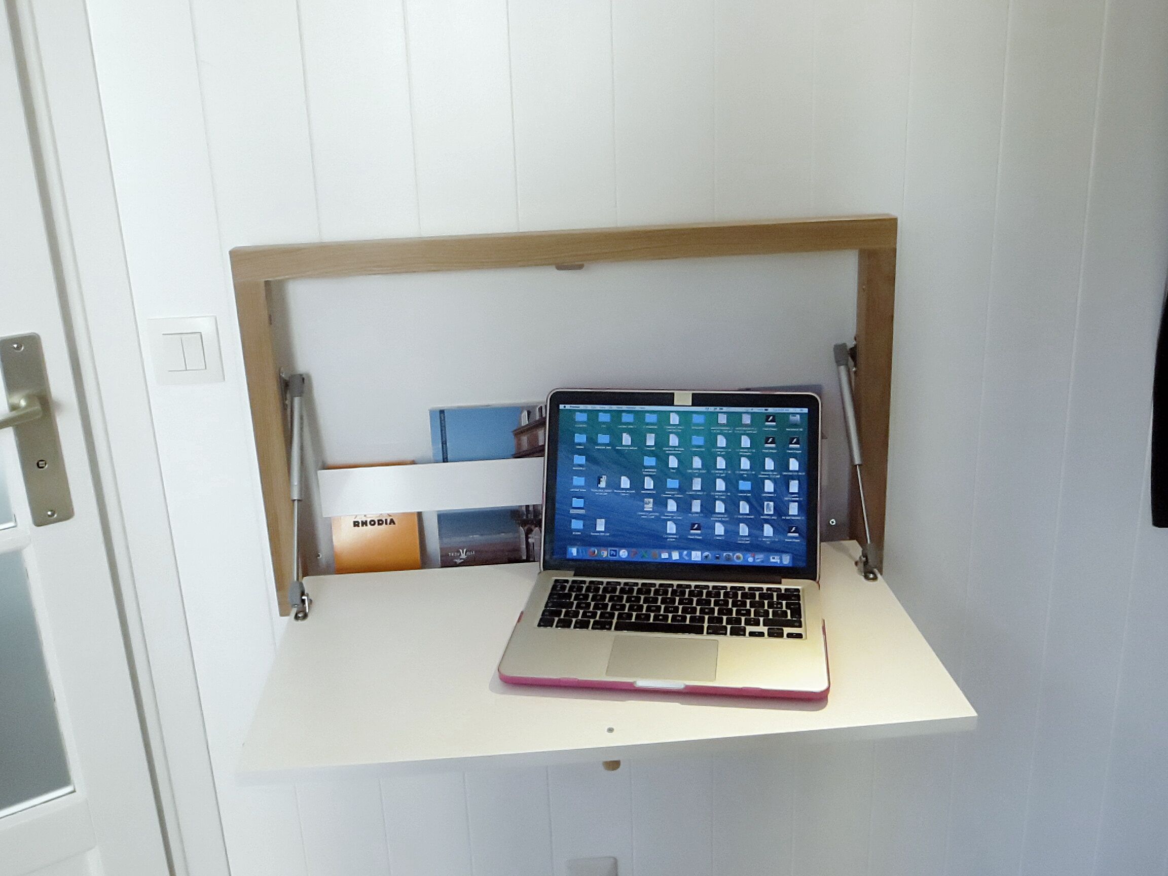 Un ordinateur portable est posé sur une étagère blanche dont le cadre est en bois, devant une porte blanche et un mur blanc.