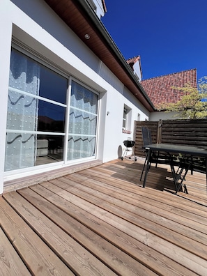 Una casa moderna con un amplio ventanal corredera, una terraza de madera y una mesa exterior negra.