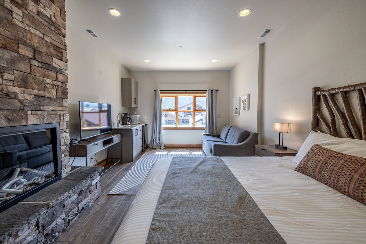 A modern hotel room with a stone fireplace, a bed, a desk, and a view of the outside.