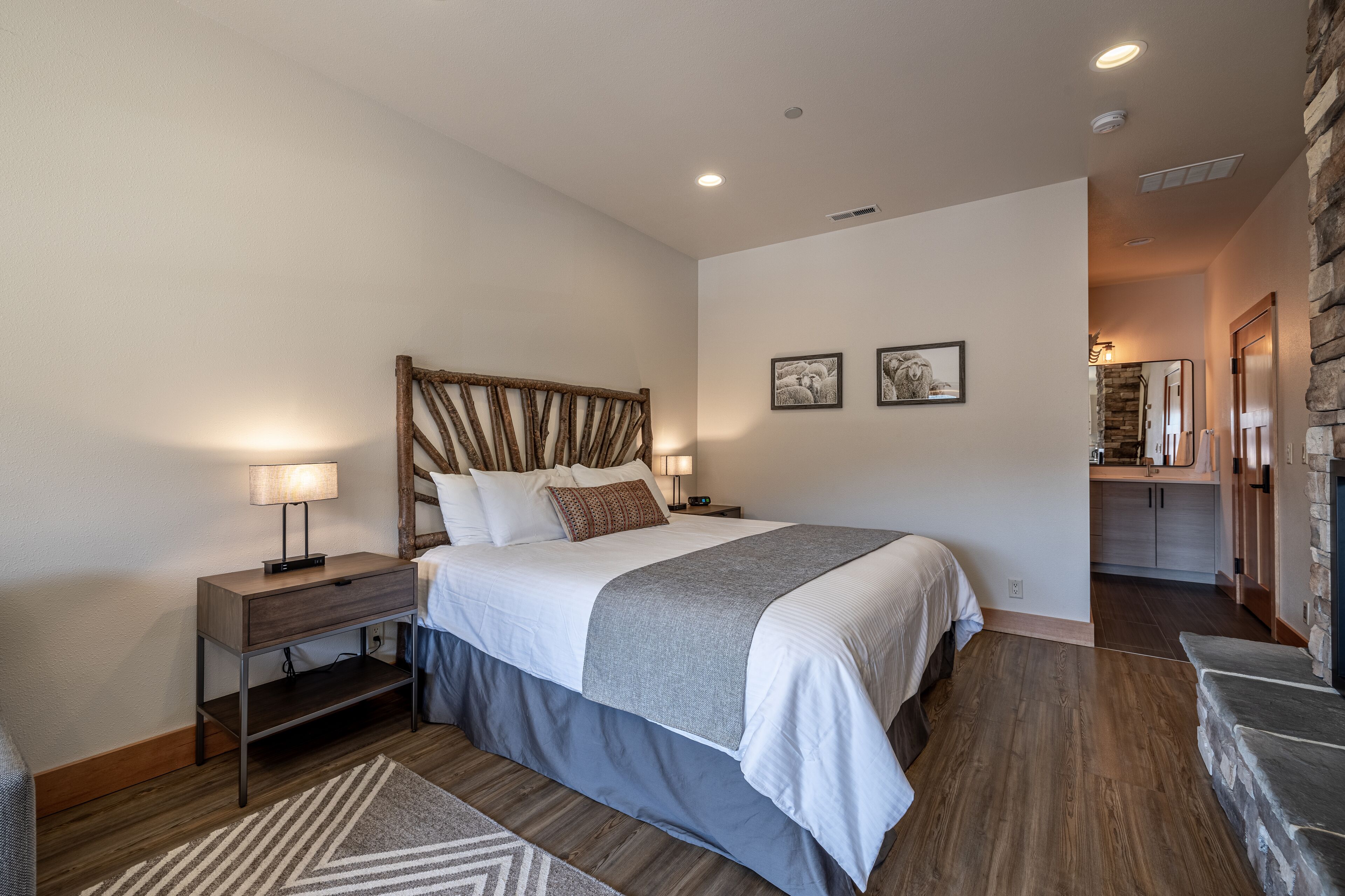 Une chambre moderne avec un grand lit, des tables de chevet, une tête de lit en bois et, au fond, une petite kitchenette.