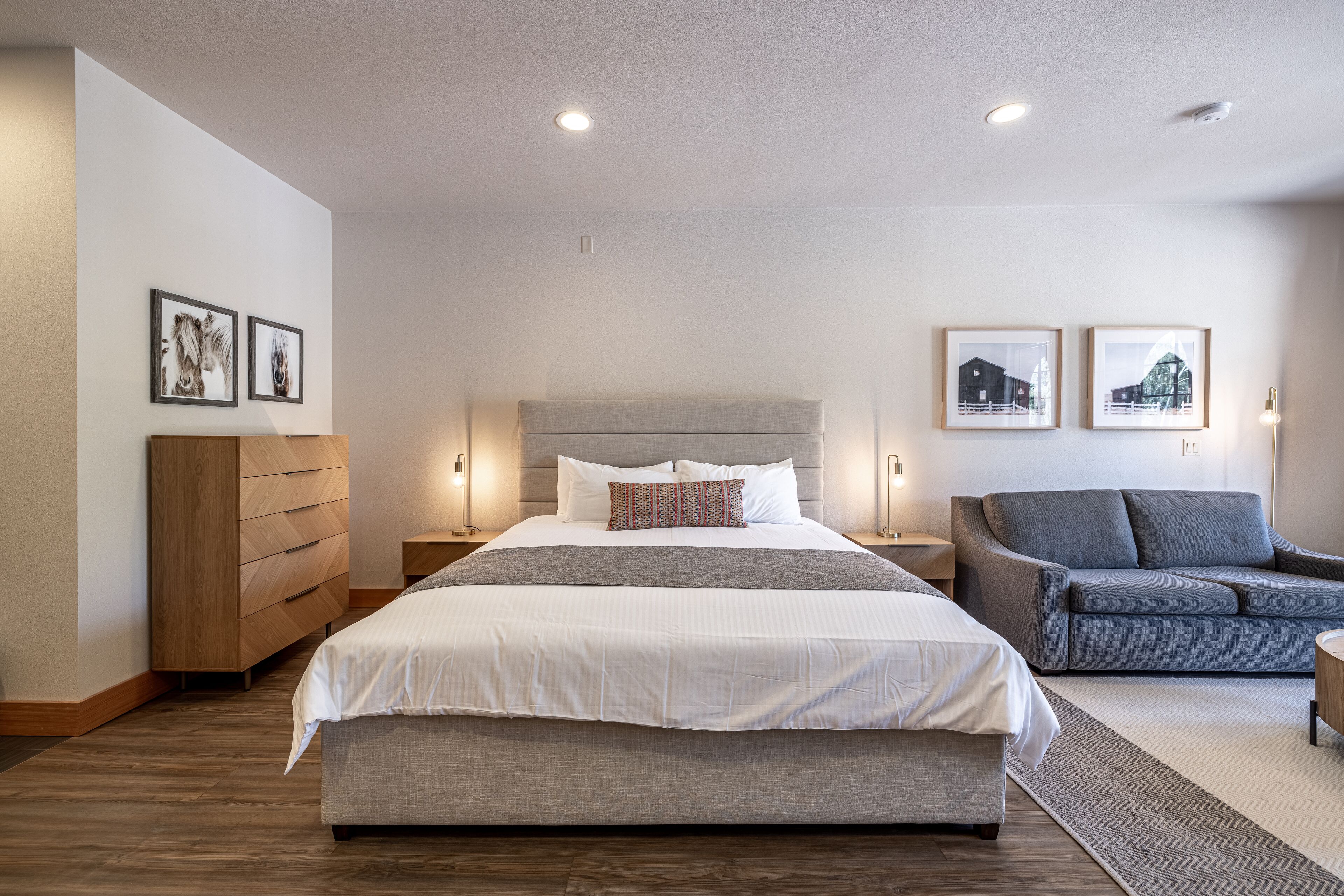 A modern bedroom with a large bed, a grey sofa, wooden furniture, and framed pictures on the walls.