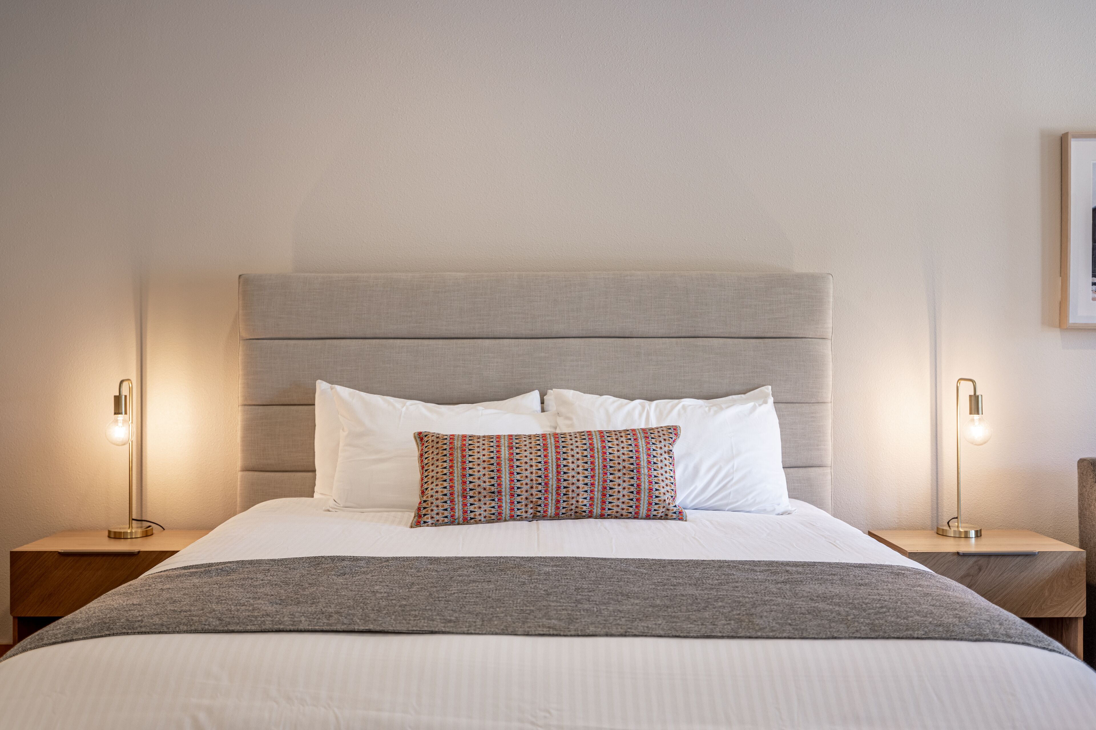 A neatly made bed with a grey headboard, white bedding, and a patterned pillow. There are two bedside tables with lamps on either side of the bed.