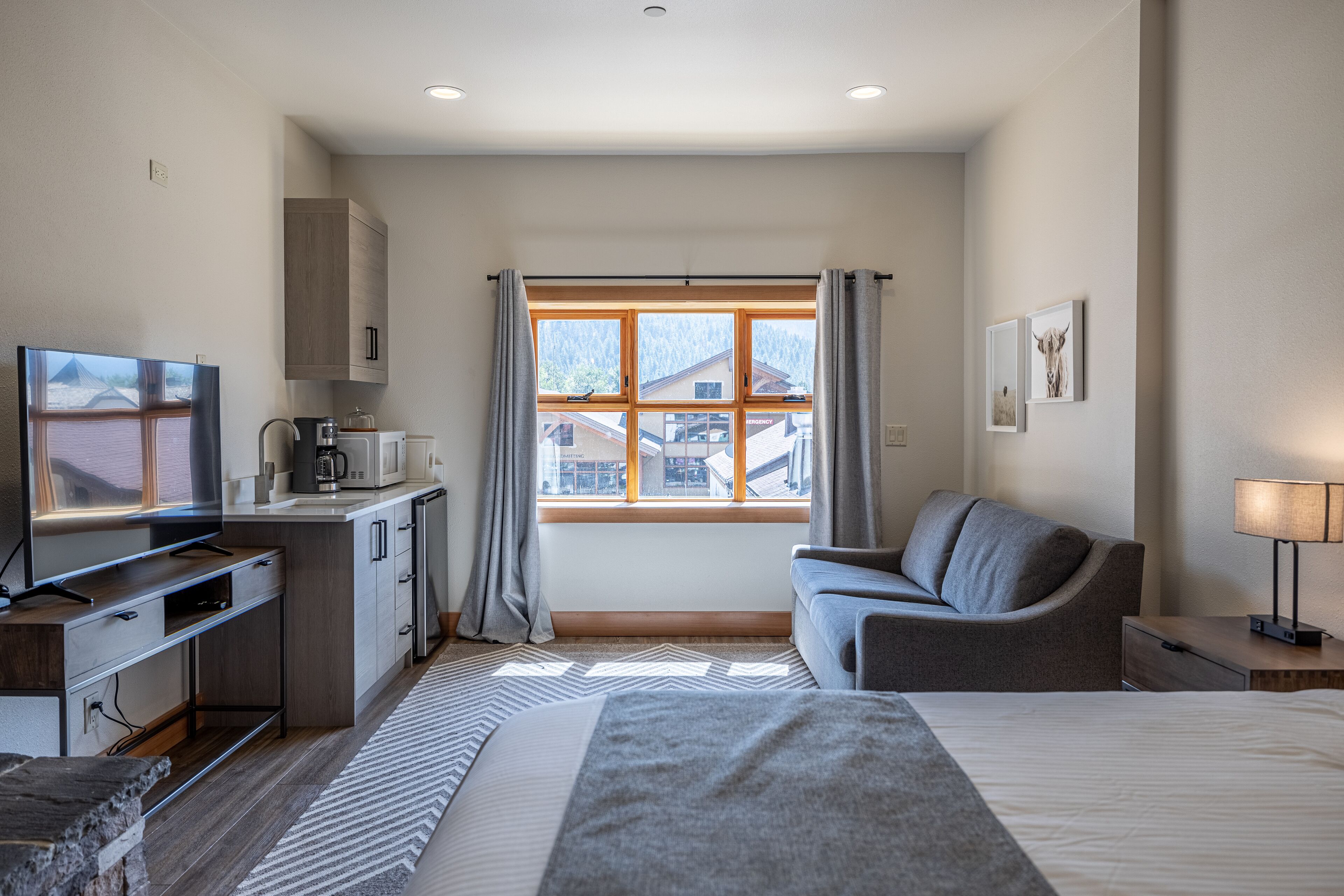 A modern hotel room with a bed, a TV, a kitchenette, and a view of the outside.
