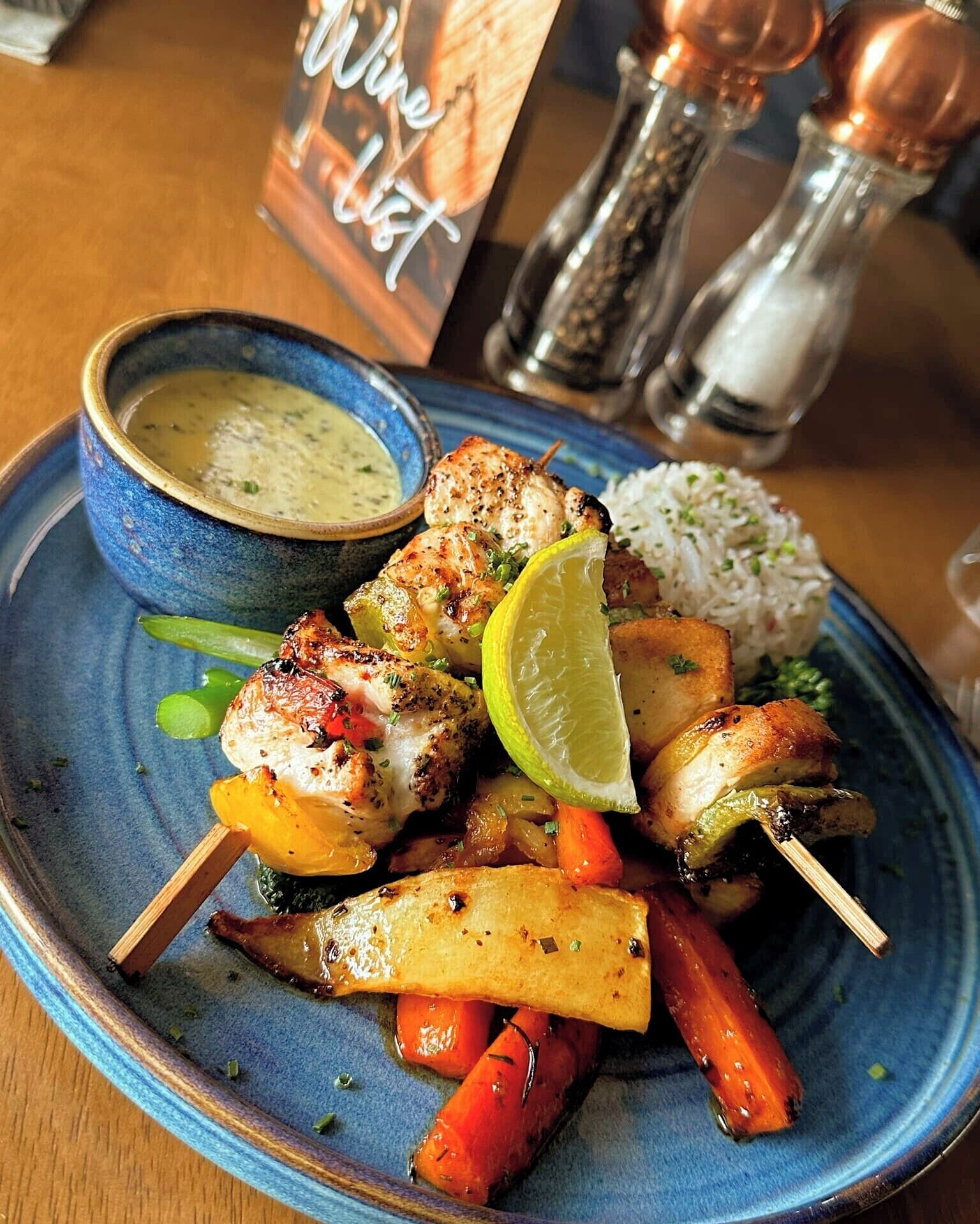 Un plato de brochetas de pollo a la parrilla con verduras y arroz, acompañado de un tazón de salsa.