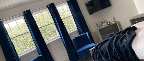 A hotel room with a bed, two blue chairs, a TV, and a window with curtains.
