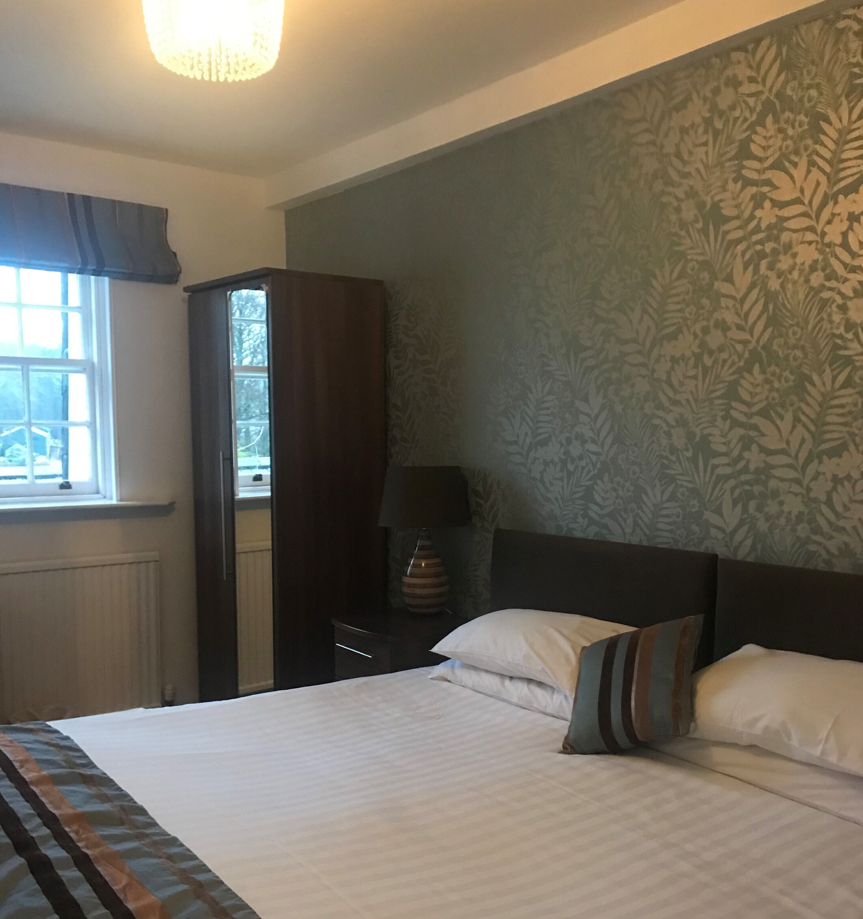 Une chambre à coucher comprenant un lit, une table de chevet, une lampe, une armoire et une fenêtre donnant sur un bâtiment à l’extérieur.