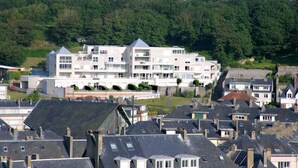 Ett hotell med flera våningar och ett platt tak, omgivet av andra byggnader, med grönskande kullar som bakgrund.