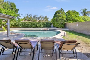 A backyard with a swimming pool, four lounge chairs, and a covered patio area.