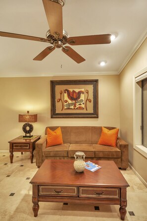 A living room with a ceiling fan, a brown sofa with orange cushions, a wooden coffee table, a side table with a lamp, and a framed abstract painting on the wall.