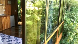 Una habitación con piso de madera, una cama con colcha estampada y vistas a un jardín exuberante a través de amplias ventanas.