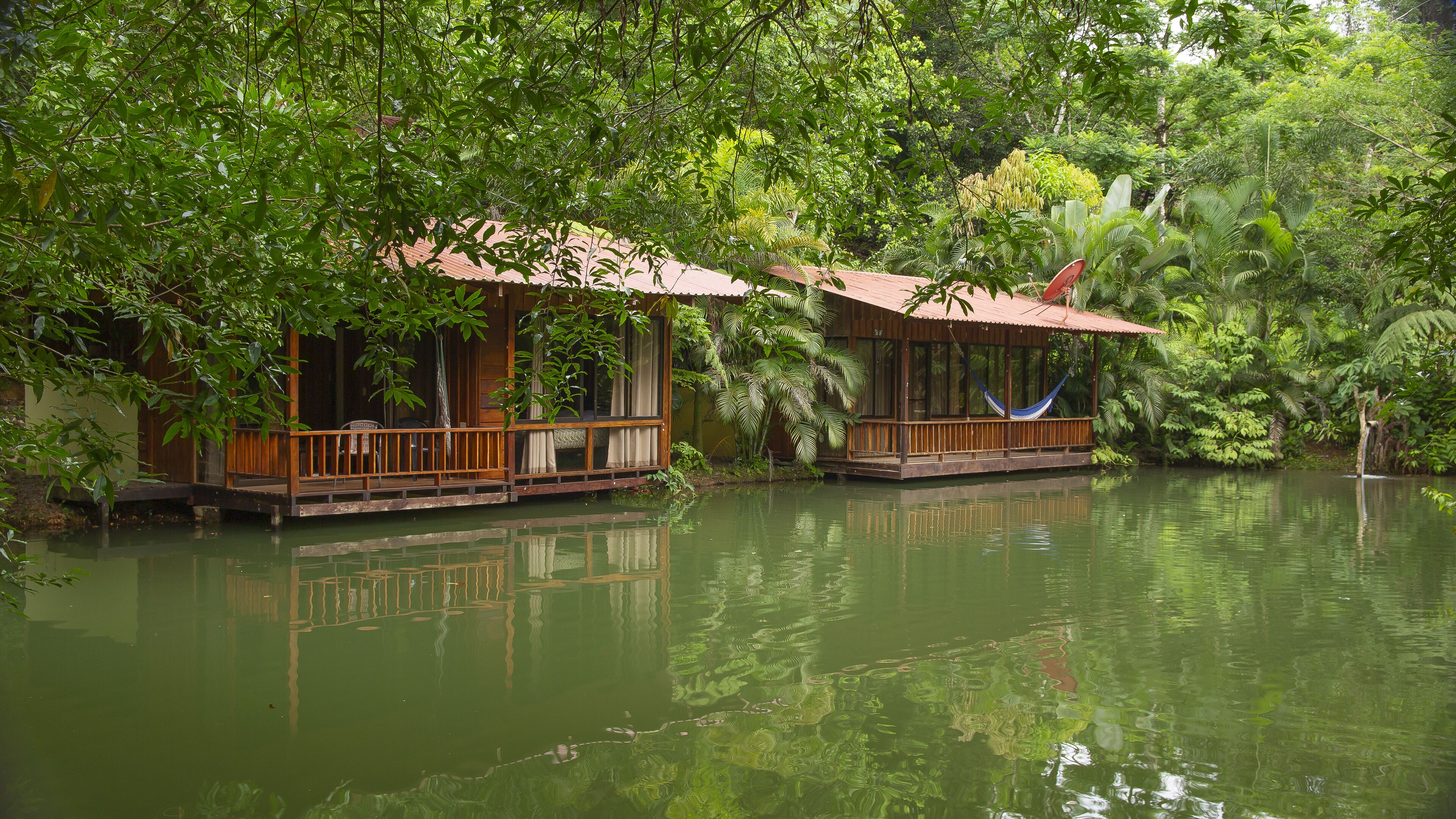 Deux maisons en bois flottantes, dotées de terrasses ouvertes, entourées d’une végétation luxuriante et bordées d’une étendue d’eau calme.