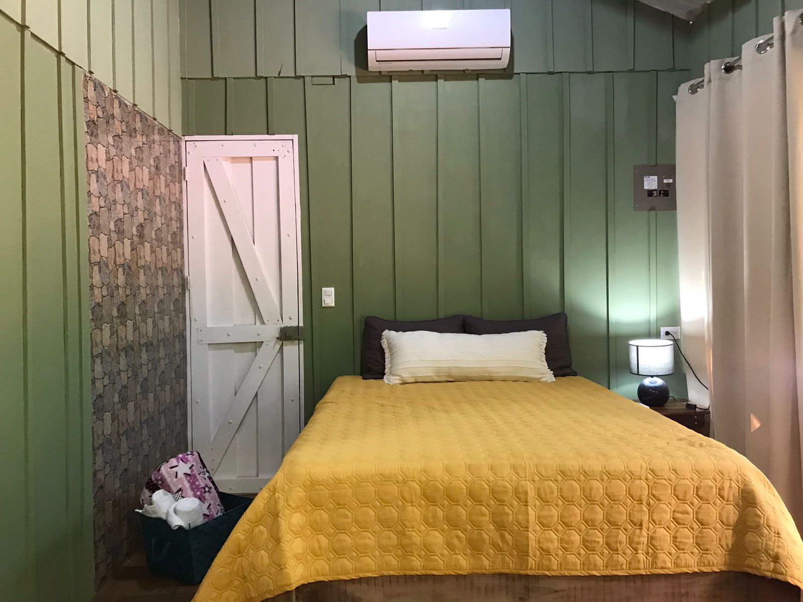 Una habitación con cama, mesita de noche, aire acondicionado instalado en la pared y puerta con textura similar a la piedra.