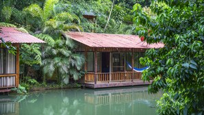 Una casa flotante de madera con un porche y una hamaca, rodeada de abundante vegetación y una masa de agua tranquila.