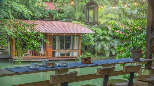 Una terraza de madera con una mesa dispuesta para el desayuno, con vistas a una casa de techo rojo y puerta de vidrio, rodeada de exuberante vegetación.