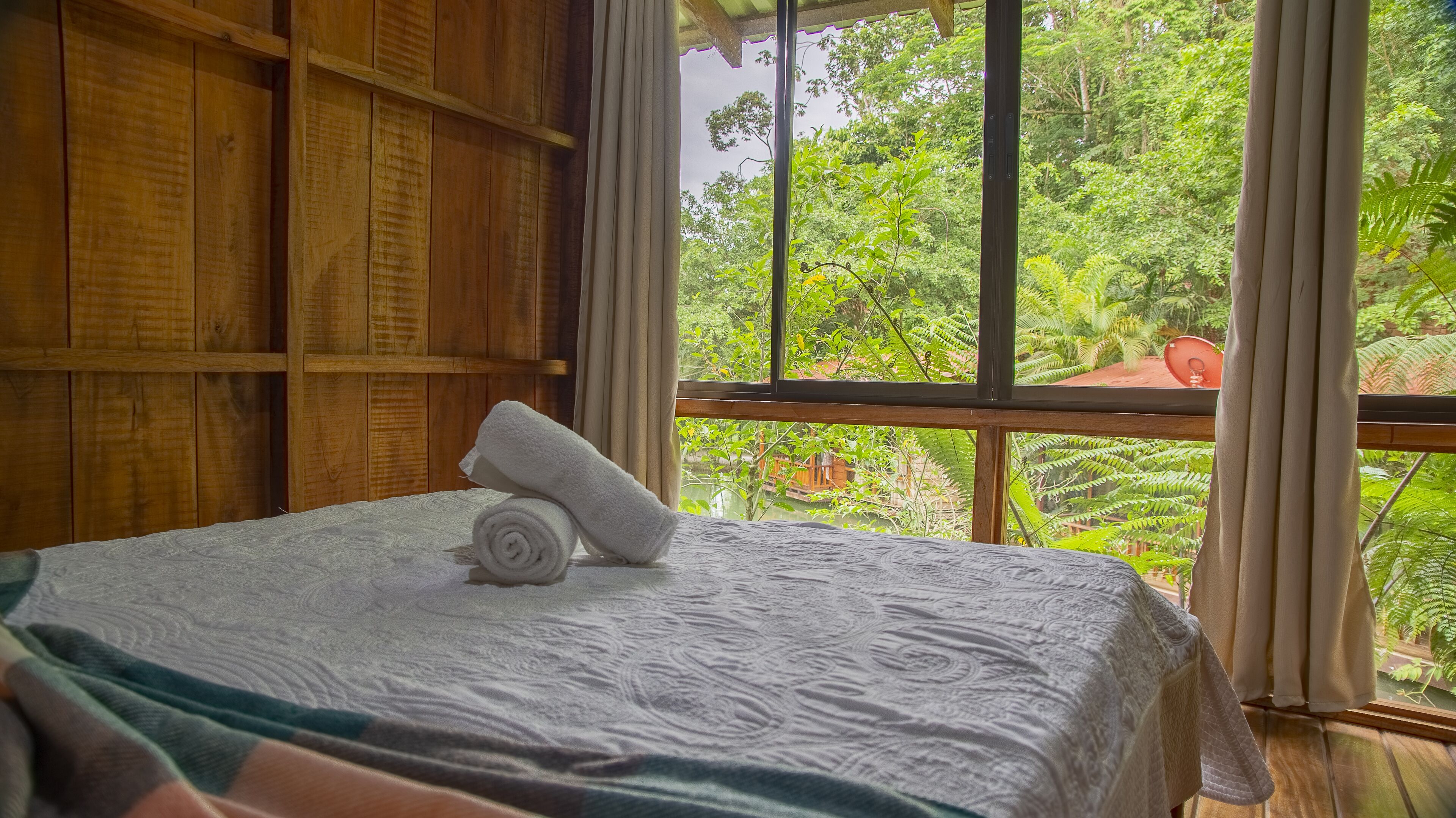 A neatly made bed with a rolled white towel, wooden paneling on the walls, and a view of lush greenery through large windows.