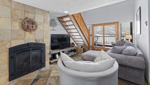 A living room with a stone fireplace, a television, a sofa, and a balcony with a view of a snowy landscape.