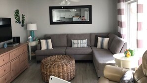 A living room with a grey sofa, a television, a ceiling fan, a mirror, and a window with curtains.
