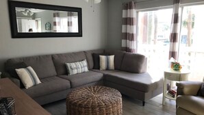 A living room with a sectional sofa, a round woven ottoman, a small side table, and a mirror on the wall.