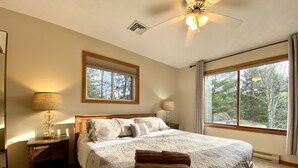 A bedroom with a large bed, two bedside tables, a ceiling fan, and a window with a view of trees. - Private 5BR Escape with Hot Tub & Games, Near Lakes, Hikes & Water Parks (Long Pond)