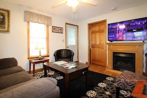 A living room with a sofa, a coffee table with books, a fireplace, and a television. - Cooperstown Baseball Rentals - Church Street House (Oneonta)