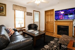 A living room with a leather sofa, a coffee table, a fireplace, and a television. - Cooperstown Baseball Rentals - Church Street House (Oneonta)