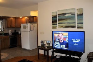 A living room with a television displaying 'The Feed' on a screen, wooden cabinets, a refrigerator, and a small table with a picture frame.