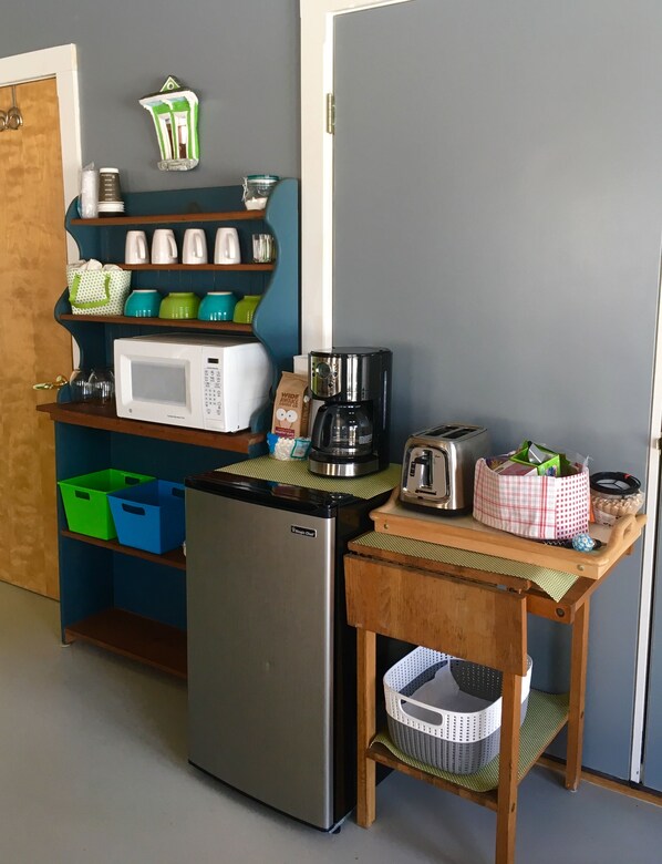 Een kleine keuken met een koelkast, magnetron, koffiezetapparaat, broodrooster en diverse keukengerei op planken.