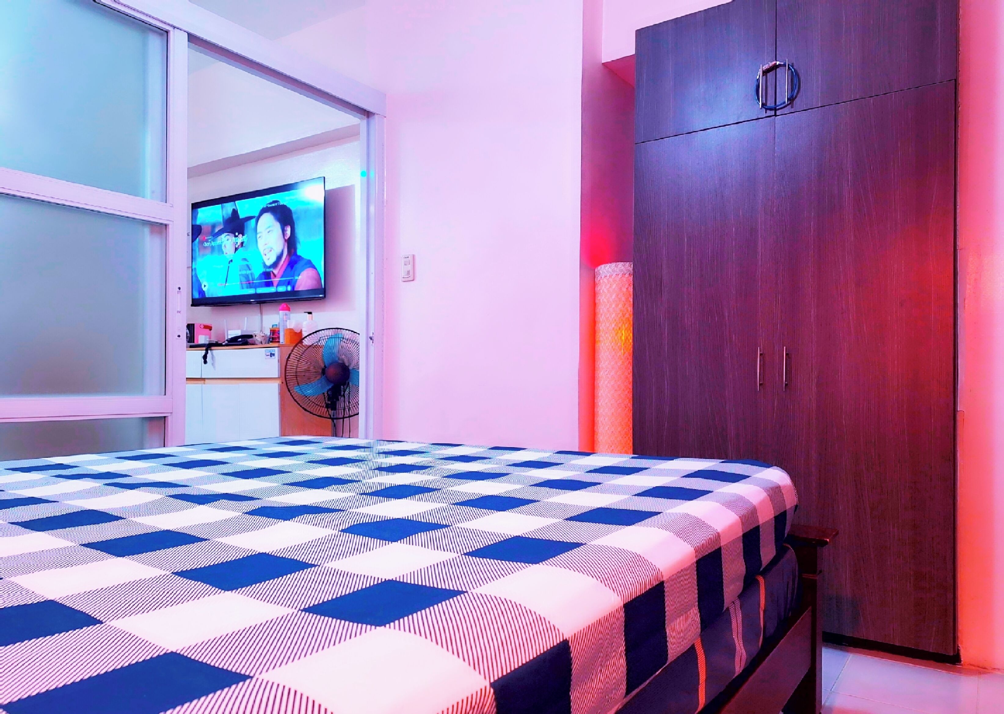 A bedroom with a checkered bedspread, a television on the wall, a wardrobe, and a fan.