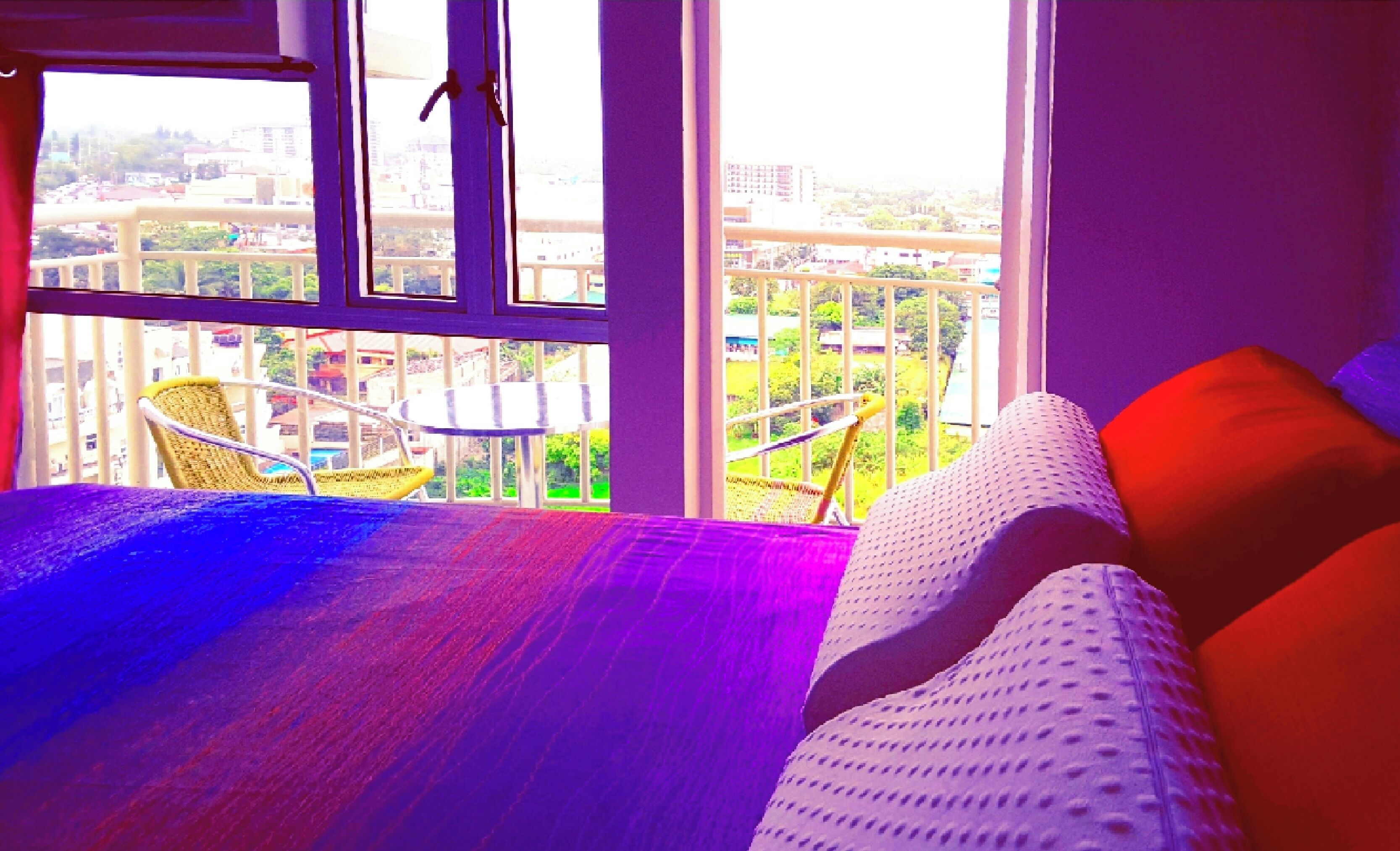 A room with a bed, a chair, and a view of a cityscape through large windows.