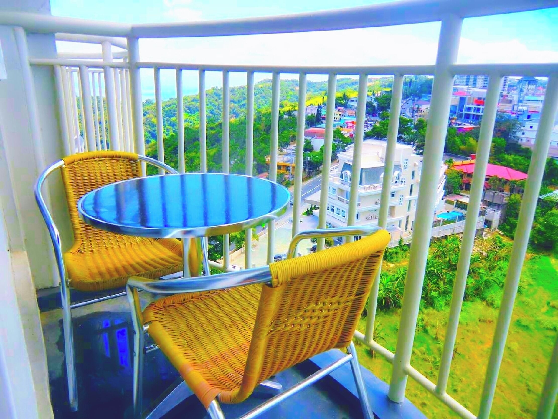 A balcony with two wicker chairs and a round table, overlooking a residential area with buildings and greenery.