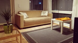 A living room with a beige sofa, a small white table, a potted plant, and a window with blinds. - The cosmopolitan apartment (Turku)