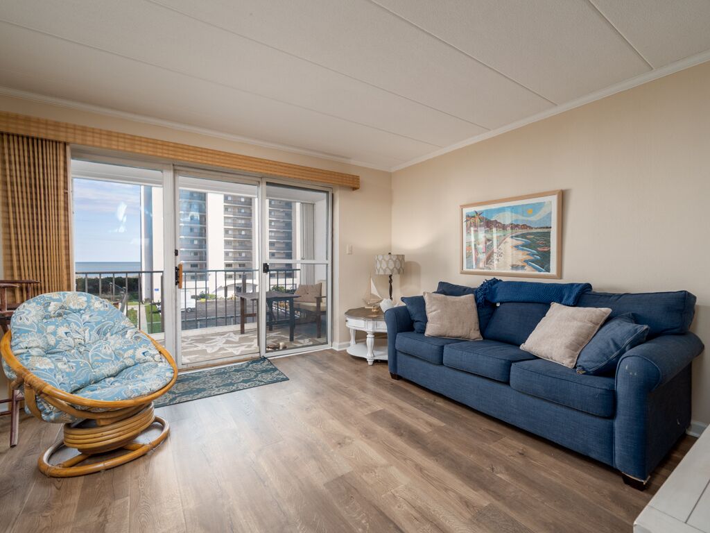 A living room with a blue sofa, a wicker chair, a glass door leading to a balcony, and a painting on the wall.