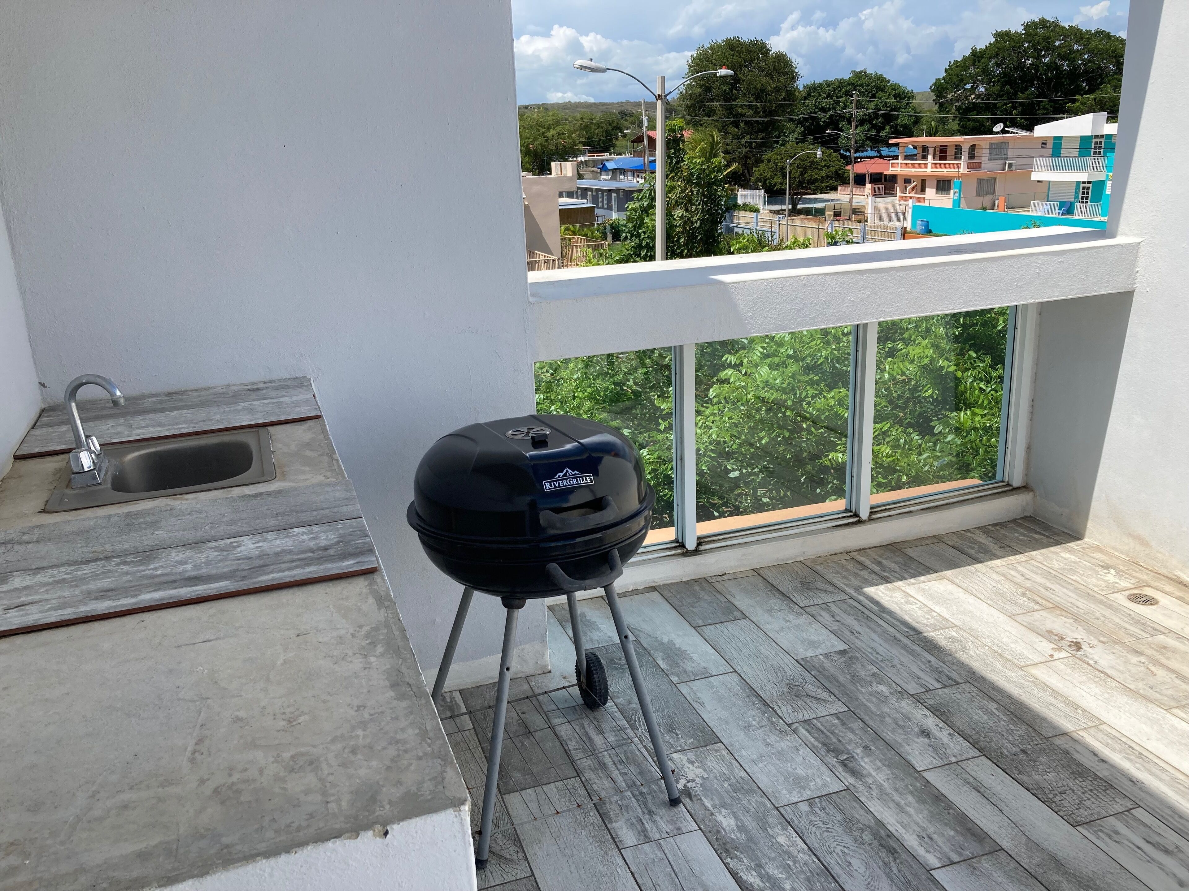 A balcony with a grill, a sink, and a view of buildings and trees.