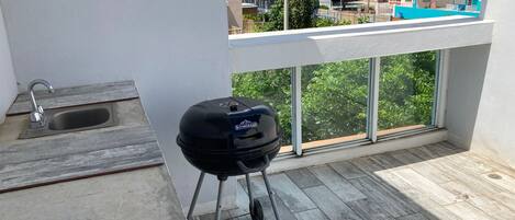 A balcony with a grill, a sink, and a view of buildings and trees.