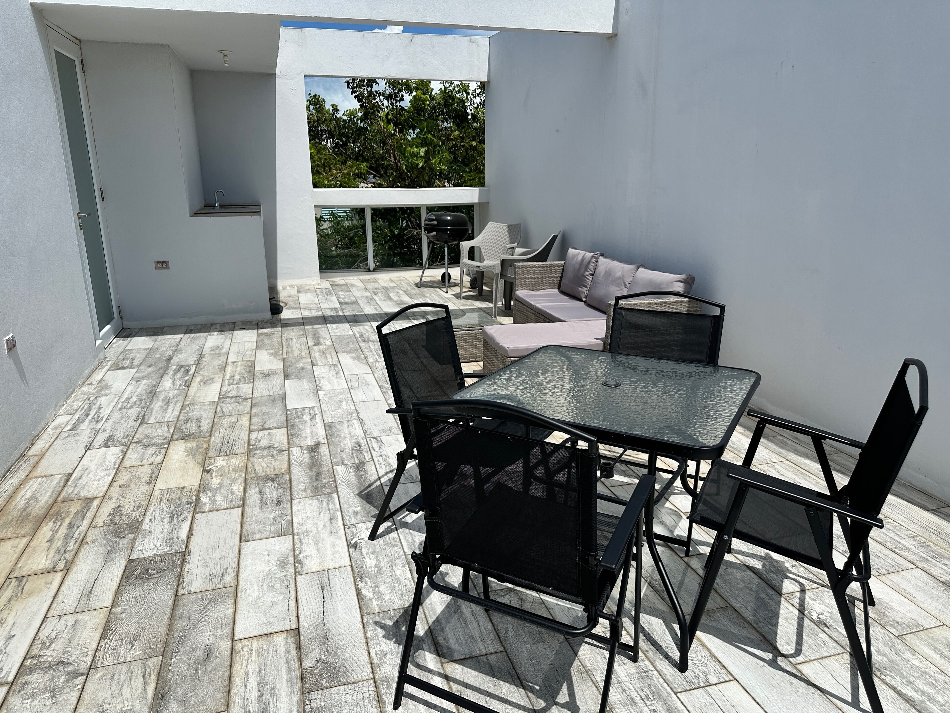 Un patio exterior con una mesa y sillas de vidrio, un sofá, y vistas a árboles a través de un ventanal grande.