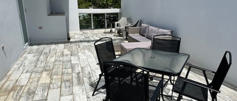 Un patio exterior con una mesa y sillas de vidrio, un sofá, y vistas a árboles a través de un ventanal grande.