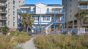 A beachfront hotel with multiple balconies, a central staircase, and a sandy pathway leading to the entrance.