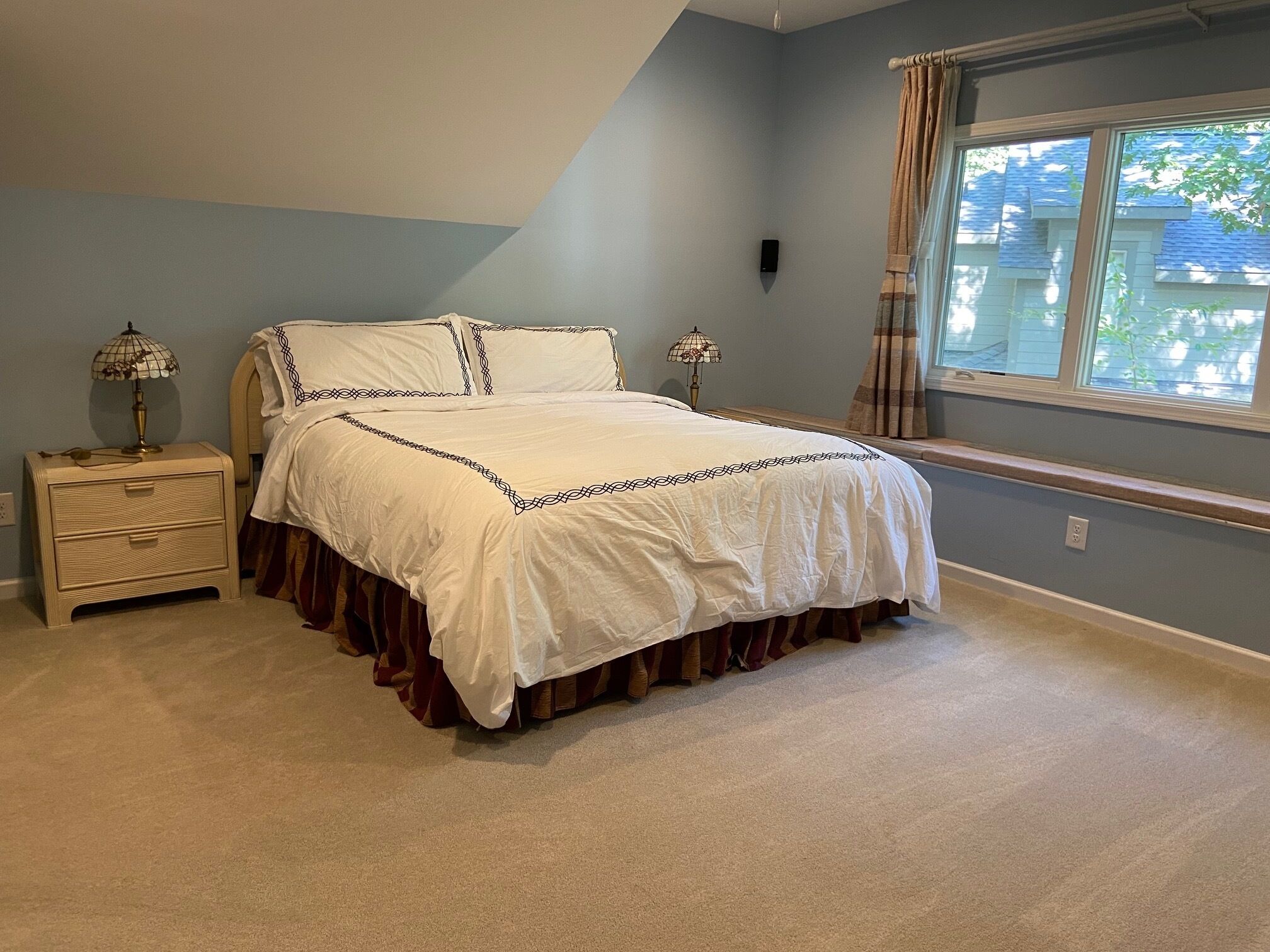 A bedroom with a bed, nightstands, a window with curtains, and a view of a house outside.
