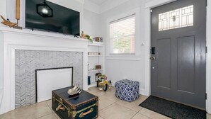 Una sala de estar con chimenea, un televisor montado encima y una puerta gris, además de un ventanal con persianas.